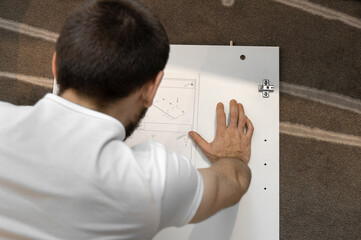 A man in a white T-shirt with a beard reads instructions for assembling a cabinet. View from the back and side. Assembling a cabinet at home. Difficulty working with furniture.