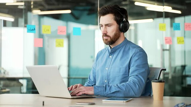 A focused IT specialist in wireless headphones works on a laptop sitting at a workplace in a business office. Serious bearded coder developer is programming, busy with a project on the computer