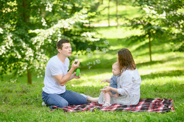 Fototapeta premium A young cheerful family with a baby is having a picnic in the park,blowing soap bubbles. Summer time concept.Family day
