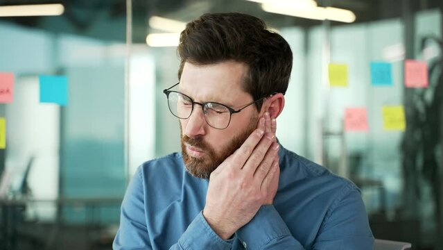 Earache. Sick businessman suffers from ear pain while sitting in business office. Upset male touches his ear with his fingers. He has furuncle, shingles, eczema, ear diseases or otitis media. Close up