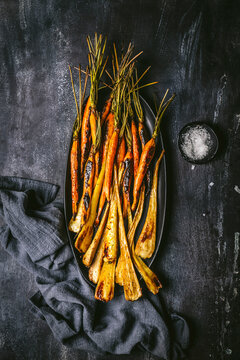 Roasted, glazed carrots and parsnips arranged on a dark oval platter with gray linen napkin on dark background