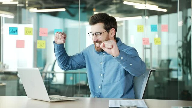 Happy handsome bearded businessman celebrating the successful completion of the project sitting at workplace in business office. Smiling satisfied crazy male employee is dancing funny. Work done
