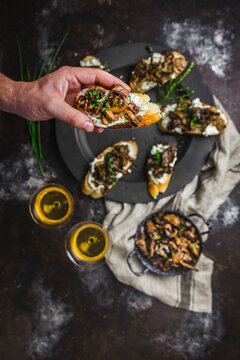 Male hand holding a Mushroom and goat cheese crostini arranged on a pewter plate, with sherry in glasses, mushrooms in small dish
