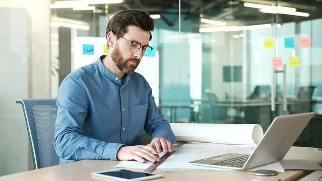 A focused handsome architect is working drawing a project on paper while sitting at desk at a workplace in a business office. Serious designer or engineer uses a pencil and a ruler to develop an idea