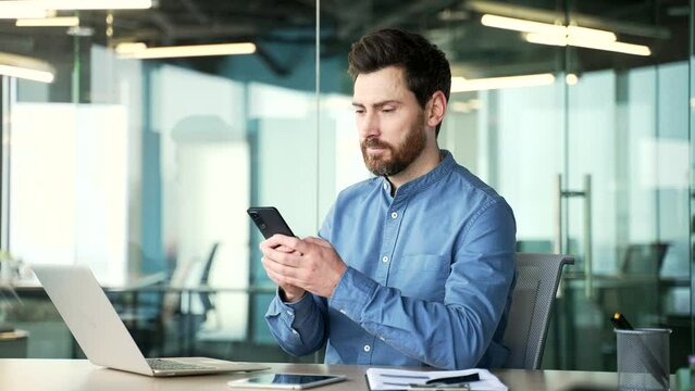 Confident bearded businessman is using phone sitting at workplace in business office. Manager works on mobile application, reading typing a message, chatting with a client, browsing or shopping online