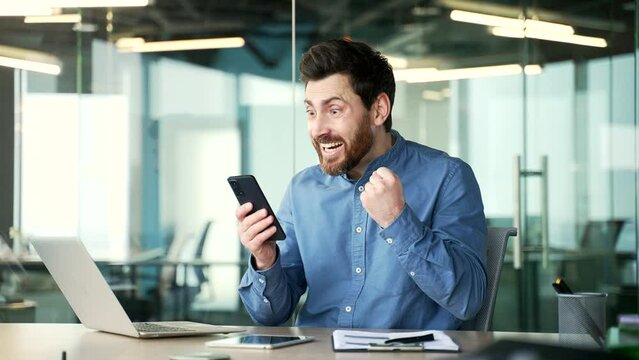 Happy handsome bearded businessman received great news on phone sitting in business office. Excited smiling worker reads a pleasant message on smartphone, celebrates success, rejoices at the win