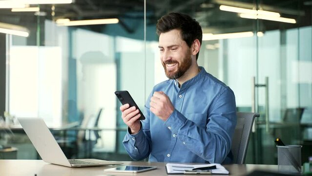 Satisfied happy handsome businessman received great news on phone while sitting at workplace in business office. Smiling glad bearded entrepreneur reads a positive good message on the smartphone