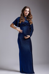 Portrait of young pretty pregnant woman on gray studio background. Female in blue sequin dress with hands near pregnant belly