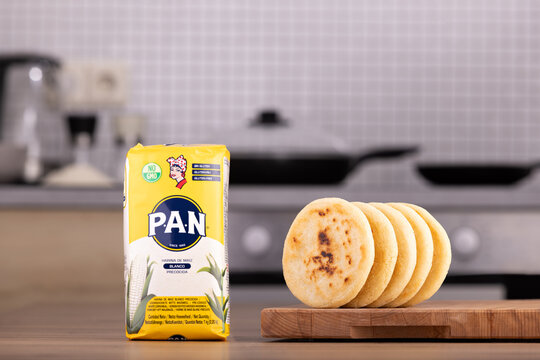 Venezuelan Harina PAN, pre-cooked white corn flour in a one kilo package on a table with some arepas cooked on one side and the kitchen as background.