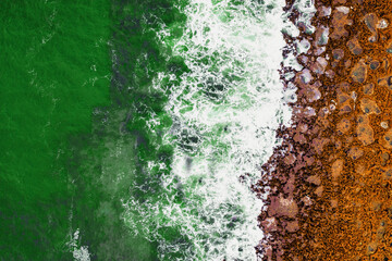 Green ocean wave, white foam and orange color rock coastline. Colors of National flag of Ireland. Aerial top down view.