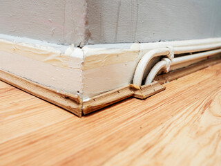 Two white water pipes attached on wooden skirting by floor of a room. Poor quality job. Poor old home system. Grey wall with trace of water leak.