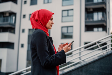 Portrait of a young happy Muslim business woman in hijab using a phone mobile near the office building.