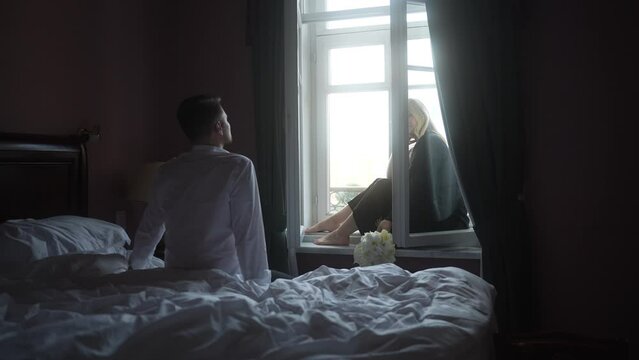 The guy looks at his woman who is sitting on the windowsill