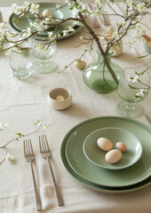Beautiful light green and beige colors morning table decoration lace tablecloth and napkins, spring cherry blossom branch, silver tableware and natural porcelain plates. Breakfast eggs healthy eating.
