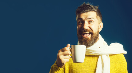 Happy man with beard in yellow sweater wrapped in white scarf with cup of hot coffee or tea. Handsome bearded man with stylish hairstyle drinking beverage in cafe, bistro. Copy space for advertising.