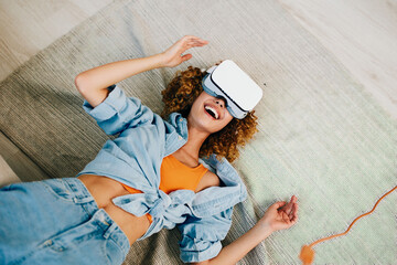 Smiling woman enjoying virtual reality game at home with futuristic VR goggles and white shirt,...