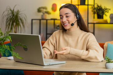 Young woman wearing headset freelance worker call center or support service operator helpline having talk with client or colleague communication support. Female girl sits at home office room at table