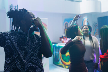 African american man singing in microphone while dancing with friends in crowded nightclub. Young clubber holding mic while clubbing partying at discotheque social gathering