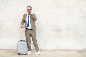 A happy senior businessman converses on his mobile phone while standing with his suitcase, demonstrating the constant connectivity of mobile professionals - obrazy, fototapety, plakaty