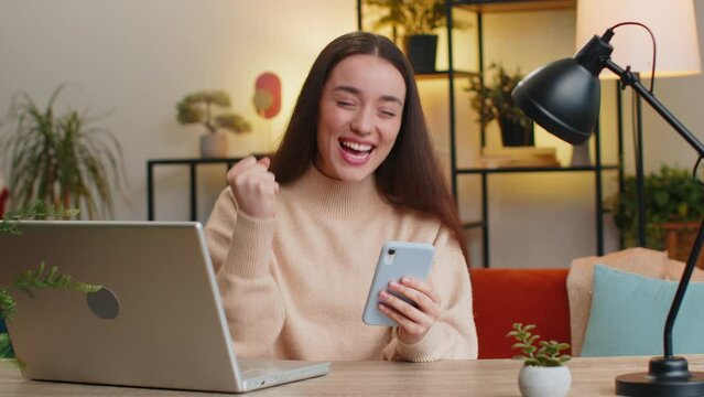 Happy woman use mobile smartphone typing browsing say Wow yes celebrating success victory winning lottery jackpot goal achievement play game good positive news triumph. Girl at home office room table
