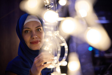In a modern restaurant ambiance, a woman in a hijab captures a selfie beside glowing lights, showcasing contemporary style and cultural diversity in a trendy urban setting.