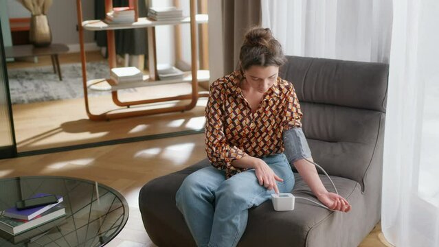 Young woman sits in modern living room and uses blood pressure monitor