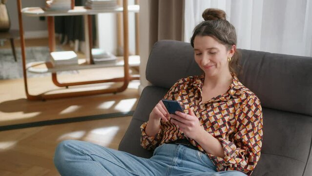 Smiling young woman sits in living room and scrolls on phone, handheld