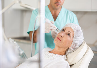 Elderly woman getting facial deep hydration procedure from experienced cosmetologist in aesthetic medicine office