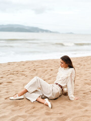 Fototapeta na wymiar Serene Summer Beauty: A Caucasian Woman Sitting on a White Sandy Beach, Enjoying the Ocean Breeze and Gazing at the Blue Sea.