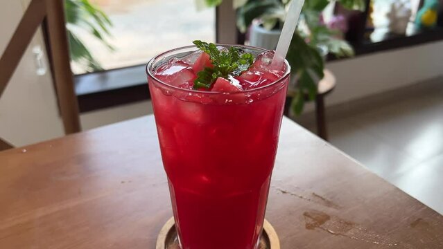 Indoor scene with red refreshing drink with ice and lime on the table Cocktail in glass in summer cafe close up. Juicy watermelon is pierced with violet straw watermelon juice