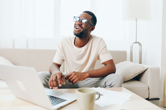 African American Man Working on Laptop with a Smile in a Modern Home Office The image depicts a young African American man sitting on a sofa, typing on his laptop in a cozy living room He looks happy