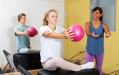 Teenager boy and girl exercising with pilates trainer latin woman in gym. They're training with pilates reformers and small fitness balls.