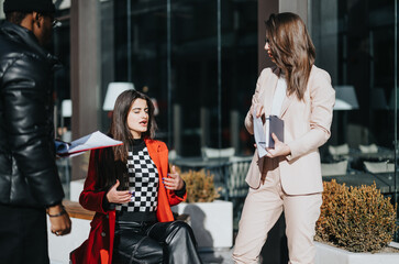 Two businesswomen and a businessman in an animated discussion, sharing paperwork on a sunny day outside modern office buildings.