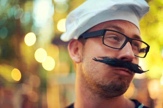 Man, fake mustache and beret in chef costume with comic face, creative and confident in french baking. Young person, glasses and cooking job in bokeh and culinary education in pastry art in europe - Powered by Adobe