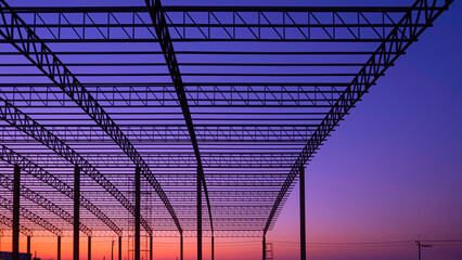 Silhouette curve metal roof beam outline with columns of large factory building structure in construction site against colorful twilight sky background, low angle view