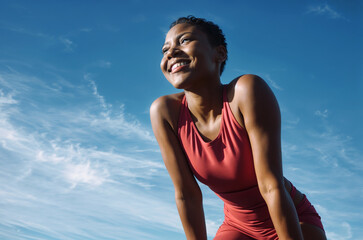 In the image, a woman is wearing a red tank top and is smiling while looking upwards. She has short hair and is crouching. The sky in the background is blue with some clouds - obrazy, fototapety, plakaty
