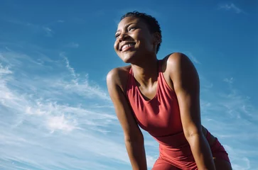 Fototapete In the image, a woman is wearing a red tank top and is smiling while looking upwards. She has short hair and is crouching. The sky in the background is blue with some clouds © Onvto