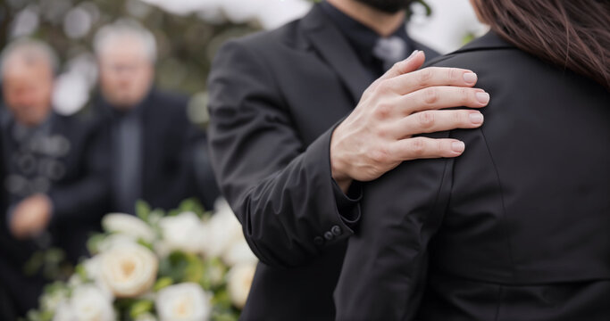 In the image, there is a funeral where people are dressed in black. A woman is hugging a man wearing a suit, and there is a close-up of their hands. A bouquet of white roses is also present