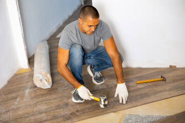 Adult man installing laminate panel on substrate with technology using soft hammer while working in...