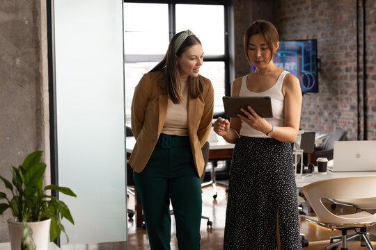 Young Caucasian woman and Asian woman discuss a tablet in a casual business office setting