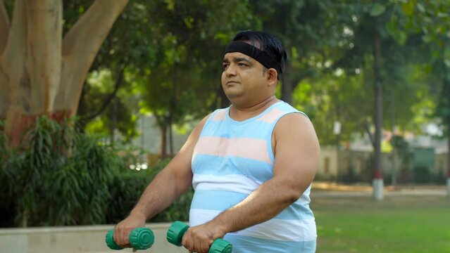 An Indian overweight man performing a workout in open gym - dumbbell lift  fitness  healthy lifestyle  obese man  BMI. A man doing a shoulder exercise with dumb bells in a park - body fitness  park...