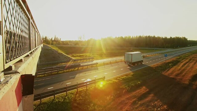 Delivery box cube van, cargo lory semi-trailer curtainsider truck driving on highway under flyover overbridge. Light truck, cars move on speedway road under overpass in countryside at sunset. Logistic