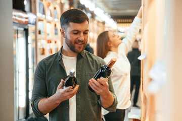 Guy holding two bottles in hands. Man and woman are choosing wine in the shop