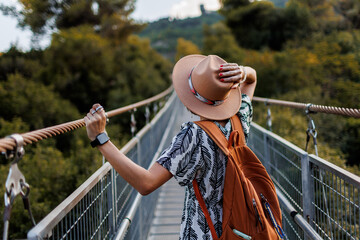 happy laughing woman in a hat walks along a pedestrian suspension bridge over the river on a sunny...