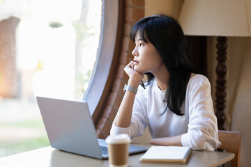 Pensive young adult woman working on a laptop in a cozy home setting, deep in thought..