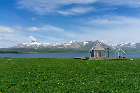  Small chapel near Paravani lake. Natural lake, blue water, clouds, snowy mountains and blue sky.