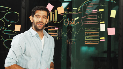 Skilled businessman looking at camera and smiling at glass wall with statistic and sticky notes....