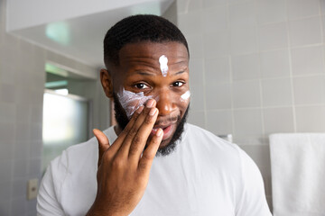 Fototapeta na wymiar Smiling black man lookin the camera and applying moisturizing cream in his bathroom