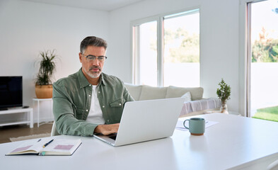 Serious older mature middle aged man wearing glasses looking at computer technology sitting at home table, using laptop hybrid working online, elearning, browsing web, searching online in living room.