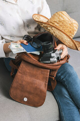 Woman packing leather backpack with retro camera, passport and dollars cash currency, straw hat,...